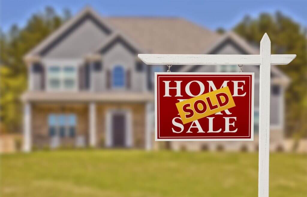 A Home For Sale sign with a "Sold" banner is in focus in front of a blurred image of a large home in the background